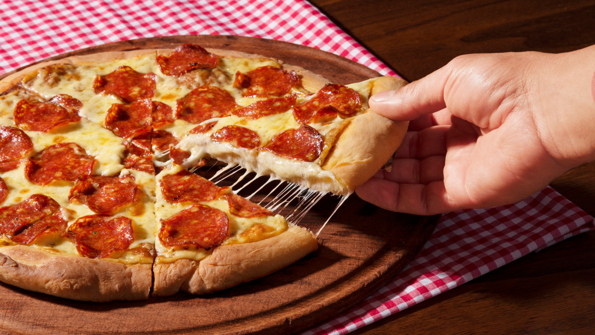 Pizza de liquidificador com calabresa em cima de um pano quadriculado vermelho e branco com uma mão pegando uma fatia da pizza
