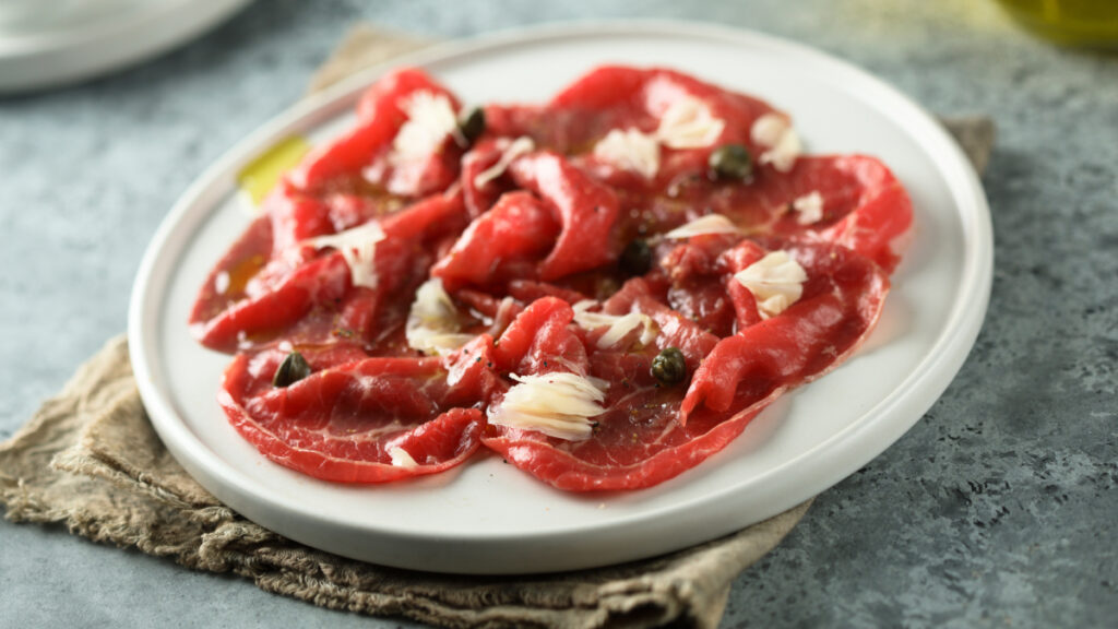 Carpaccio servido em prato branco com queijo parmesão e alcaparras