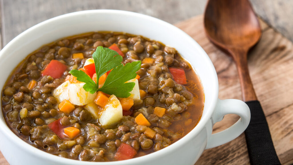 Sopa de lentilha servida em bowl branco com pedaços de tomate, cenoura, batata e uma folha verde