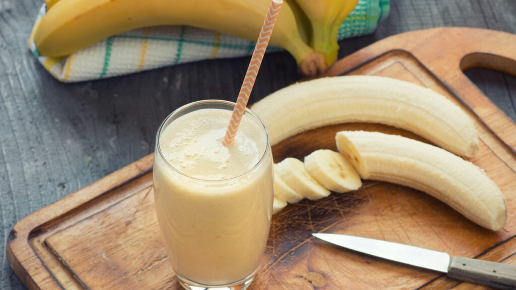 Vitamina de banana com canudo em cima de uma tábua de madeira e bananas fatiadas ao lado