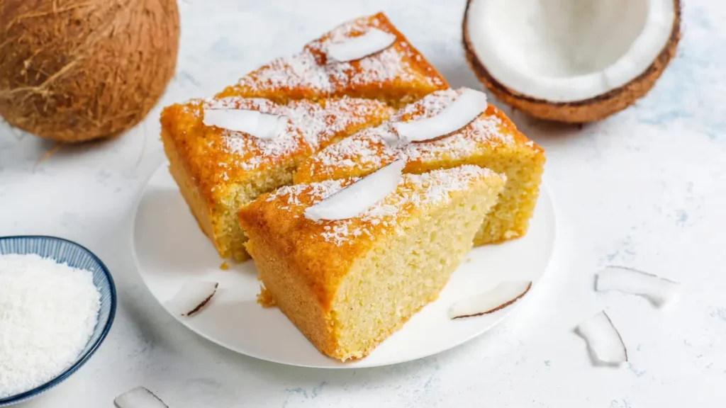 4 fatias de bolo de coco servidas em um prato branco com um coco ao lado