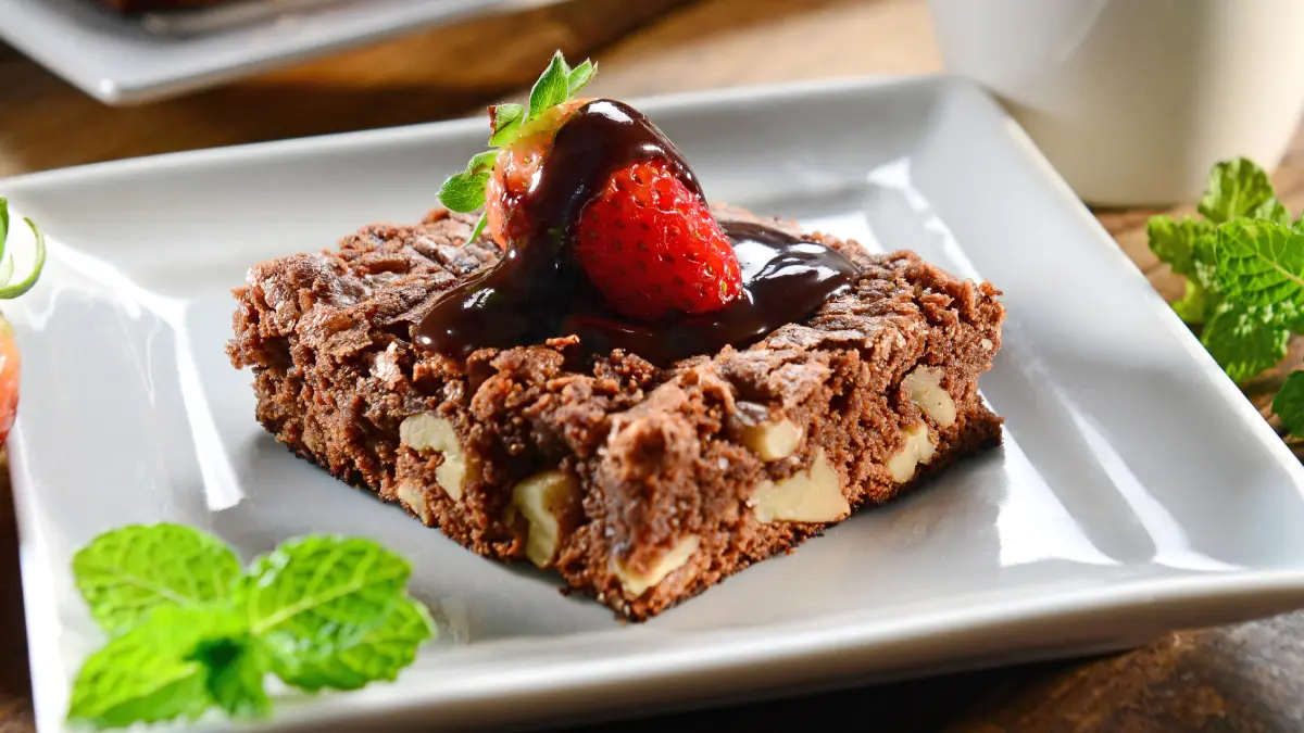 Brownie de nescau com morango e calda de chocolate servido em um prato branco quadrado