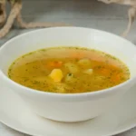 Sopa de legumes servida em bowl branco de porcenala em mesa de madeira.