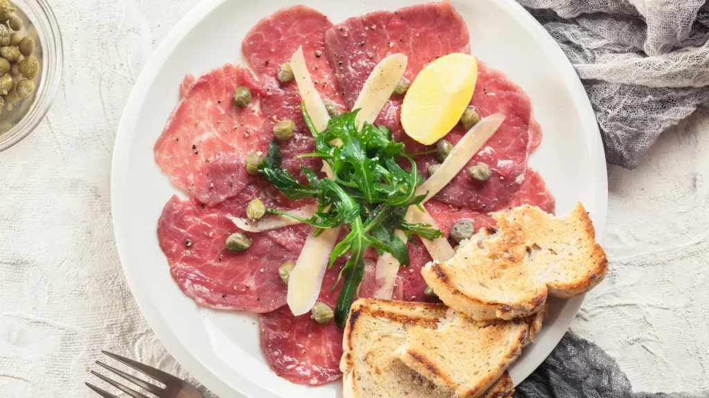 Carpaccio de carne visto de cima servido em prato branco com folhas verdes em cima