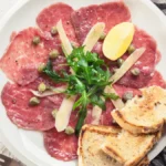 Carpaccio de carne visto de cima servido em prato branco com folhas verdes em cima