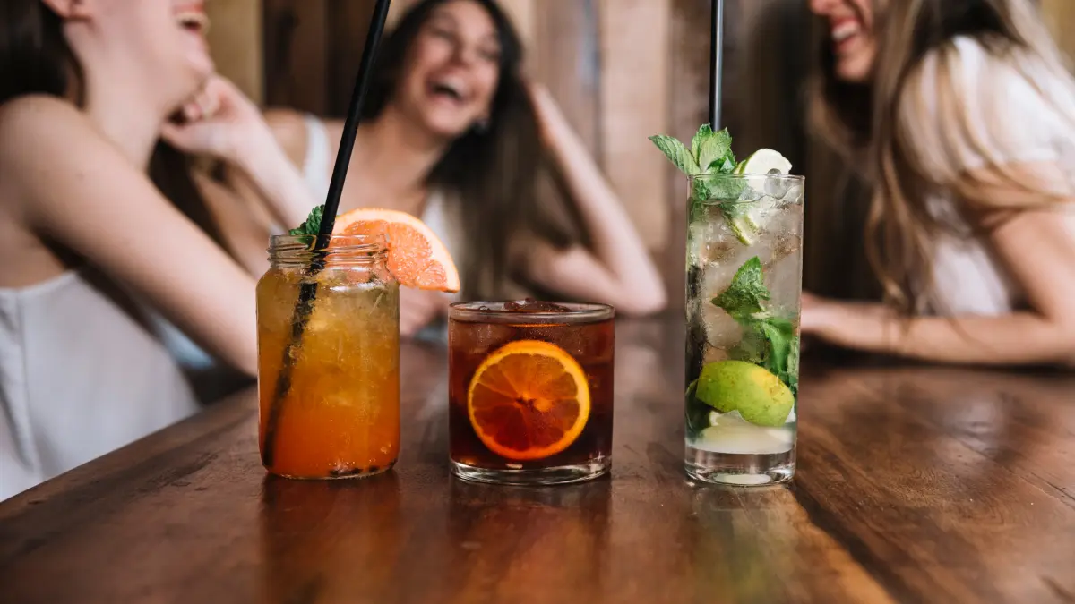Drinks com gin em cima de mesa de madeira com três mulheres sorrindo ao fundo da foto