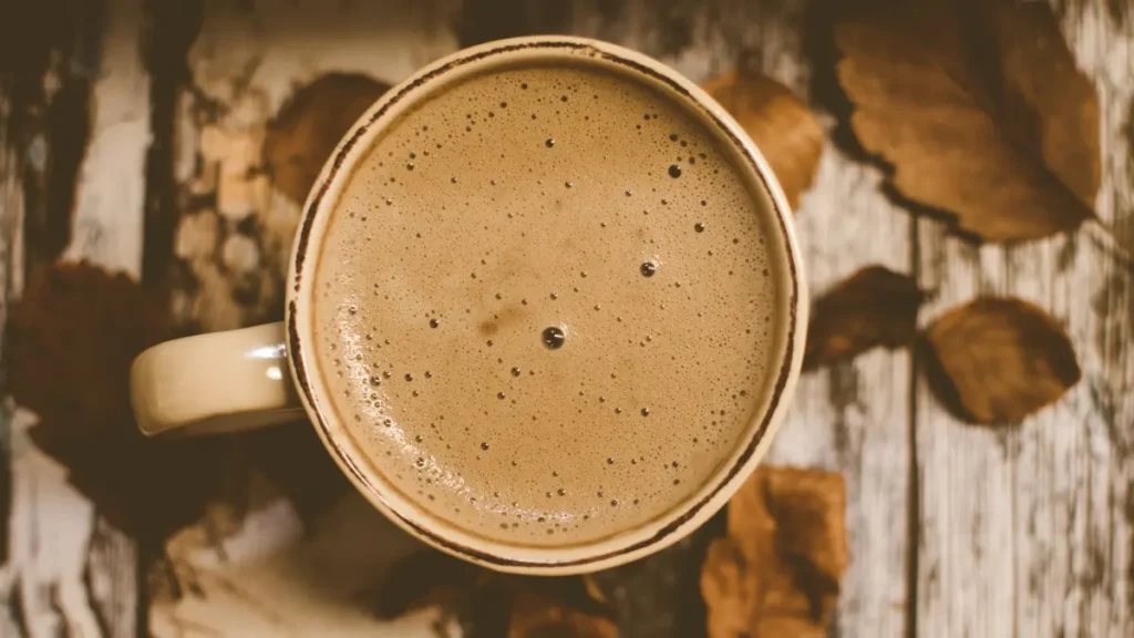 Capuccino caseiro com espuma em xícara de cerâmica com bordas arranhadas vista de cima em cima de mesa de madeira com folhas secas ao lado