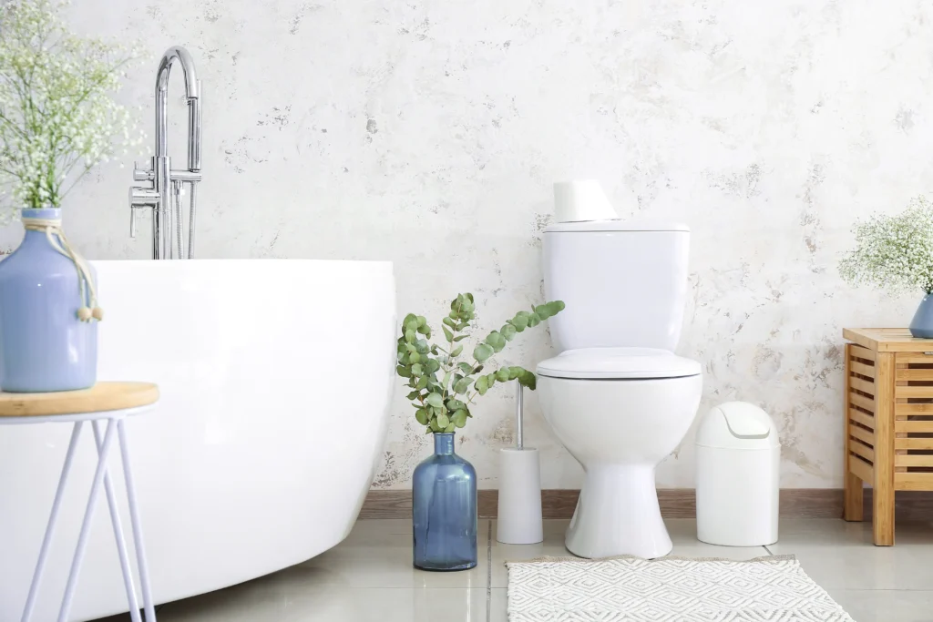 Banheiro com banheira e vaso sanitário de cor branca, revestimento marmorizado de tom claro e nuances de cinza e objetos decorativos variados espalhados pelo ambiente.