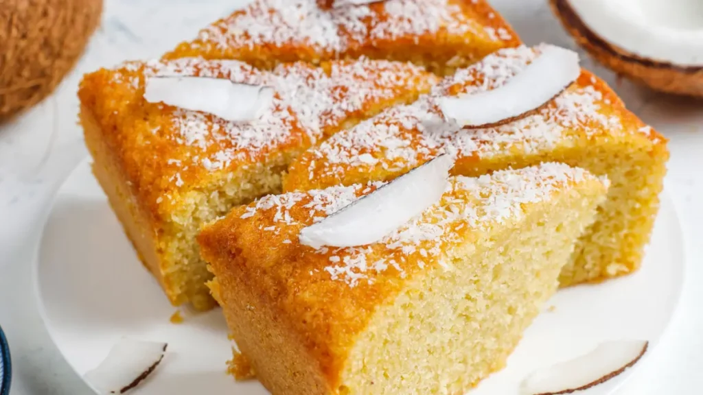 Bolo de arroz cru, fofinho, com côco ralado polvilhado por cima e decorado com lascas de côco fresco.