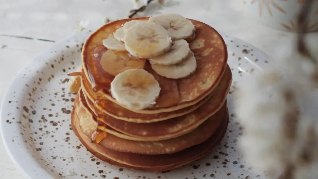 6 unidades de panqueca de banana com aveia servidas em prato branco finalizadas com bananas fatiadas e fio de mel