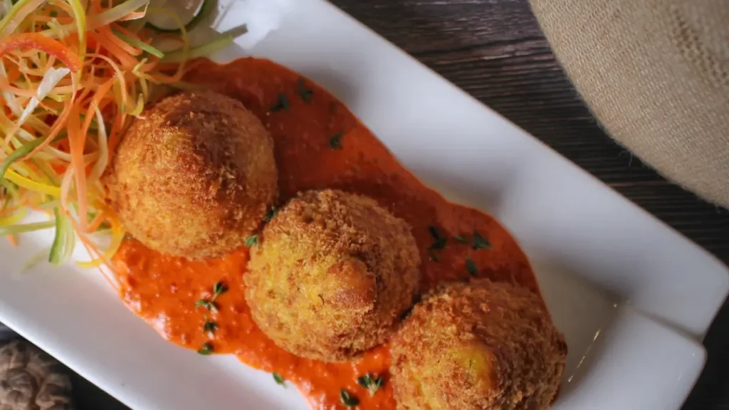 Bolinhas de arancini servidas sobre molho de tomate e acompanhamento de salada de legumes refogados