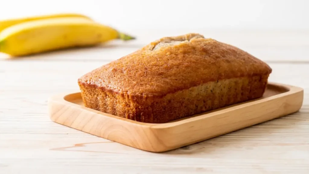 Bolo de banana de liquidificador em tábua de madeira e duas bananas no plano de fundo embaçadas