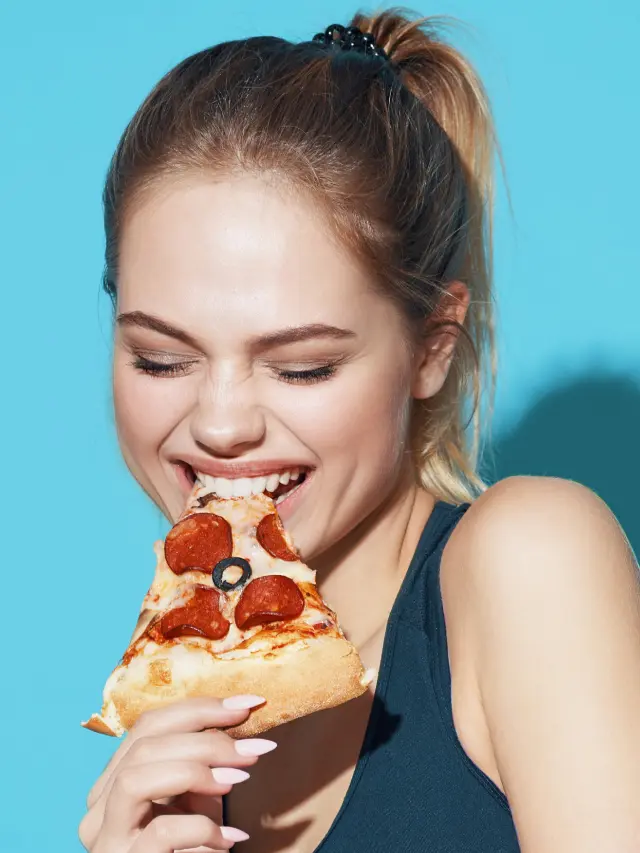 Mulher loira mordendo pedaço de pizza de calabresa e fundo azul