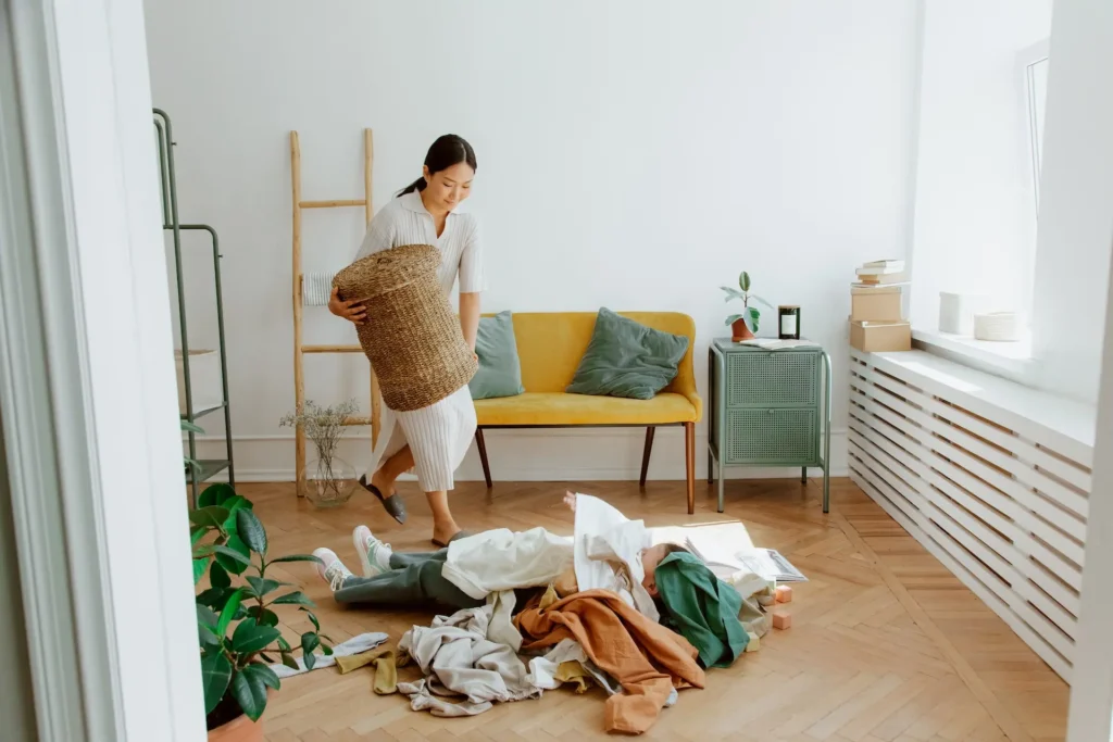 Mulher asiática carregando cesto em fibra natural, para encher com as roupas que estão espalhadas pelo chão.