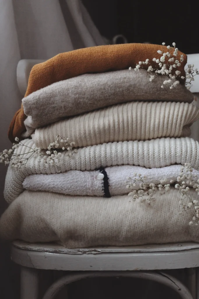 Peças de roupa em tricot, empilhadas sobre uma cadeira e flores decorando a imagem.