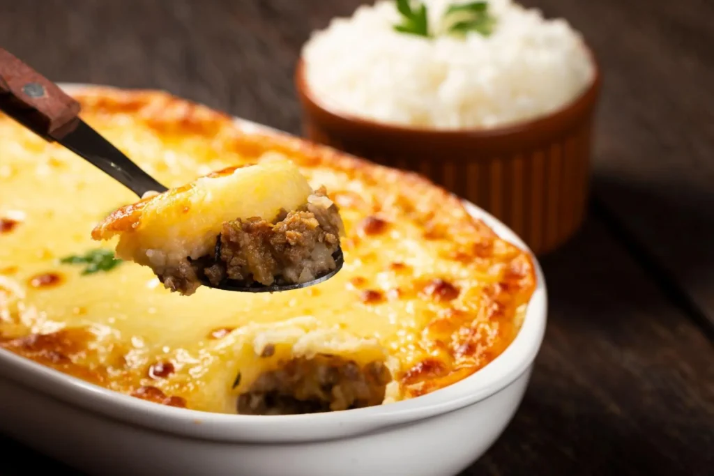 Escondidinho de mandioca com carne moída servida em travessa branca com colher pegando uma porção