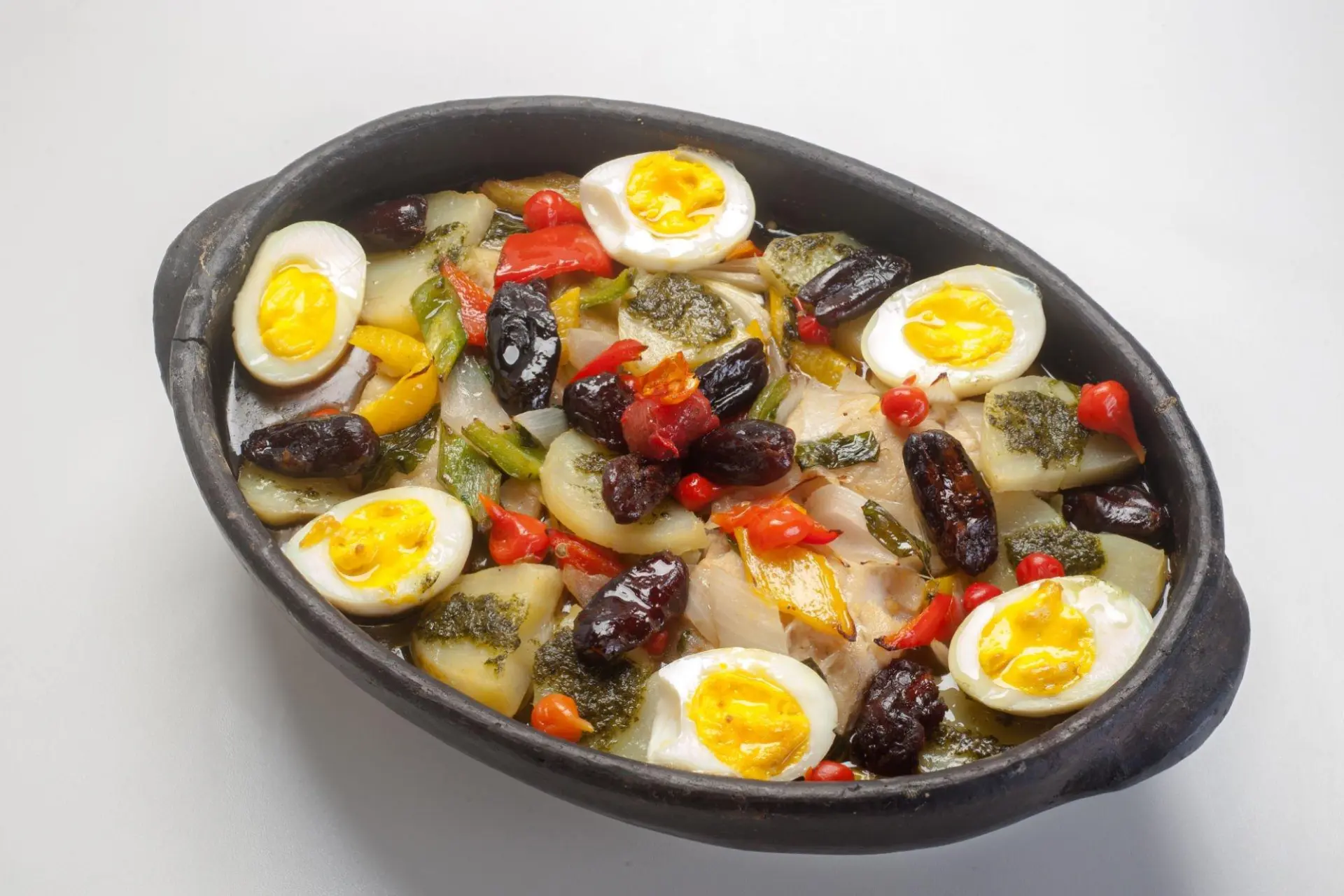 Bacalhau a gomes de sá, servido em travessa de cor escura e decorado com fatias de ovos, azeitonas, cebolas e pimentões coloridos