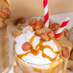 Milkshake de amendoim em copo de vidro, decorado com chantilly, calda de caramelo e grãos de amendoim. E canudo listrado de vermelho e branco.