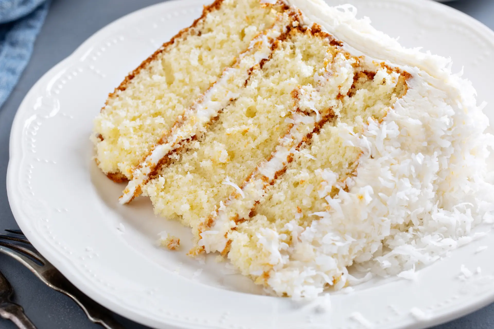 Fatia de bolo com massa branca e cobertura e recheio de prestígio com coco ralado, servido em prato branco com talheres ao redor