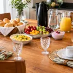 Crie uma imagem de mesa posta de café da manhã, elegante, alegre, em uma área com luz do dia, com 6 lugares montados em frente as suas respectivas cadeiras, talheres, xícaras, copos e guardanapos bem posicionados, pão francês na cesta forrada com um paninho, frutas em uma travessa, garrafa térmica de café, Jarra de água, Jarra de suco, iogurte e ovos mexidos.